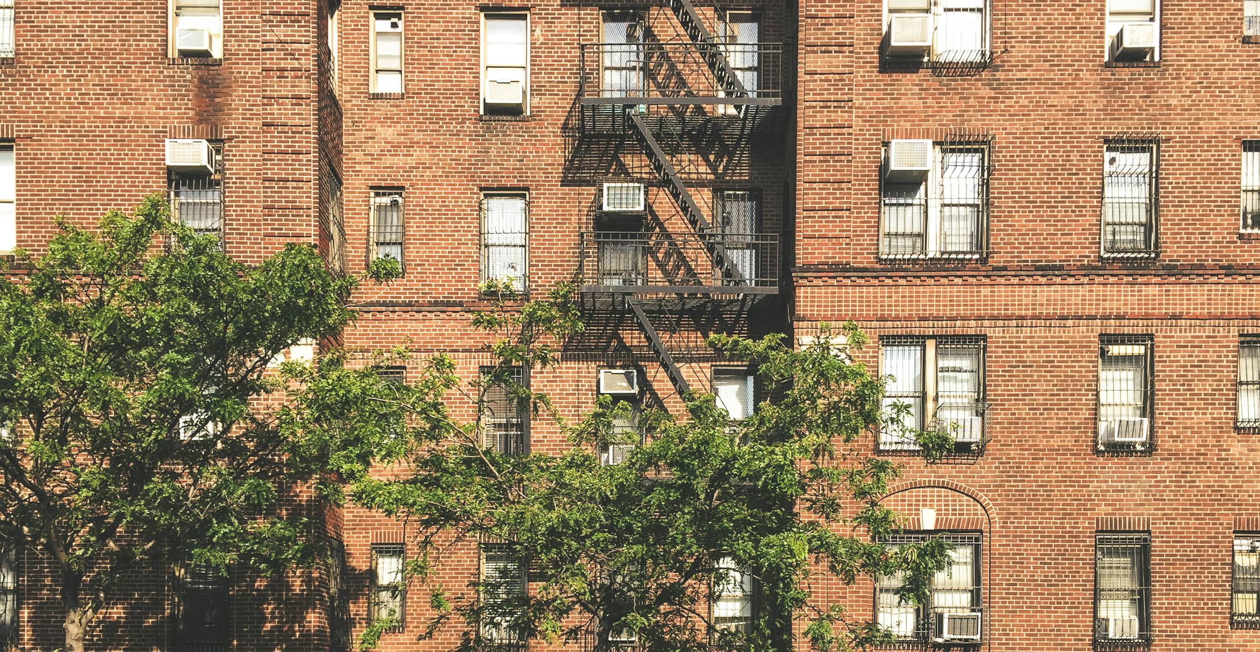 a classic brick apartment building with fire escape and air conditioners surrounded by trees. 2597999 scaled